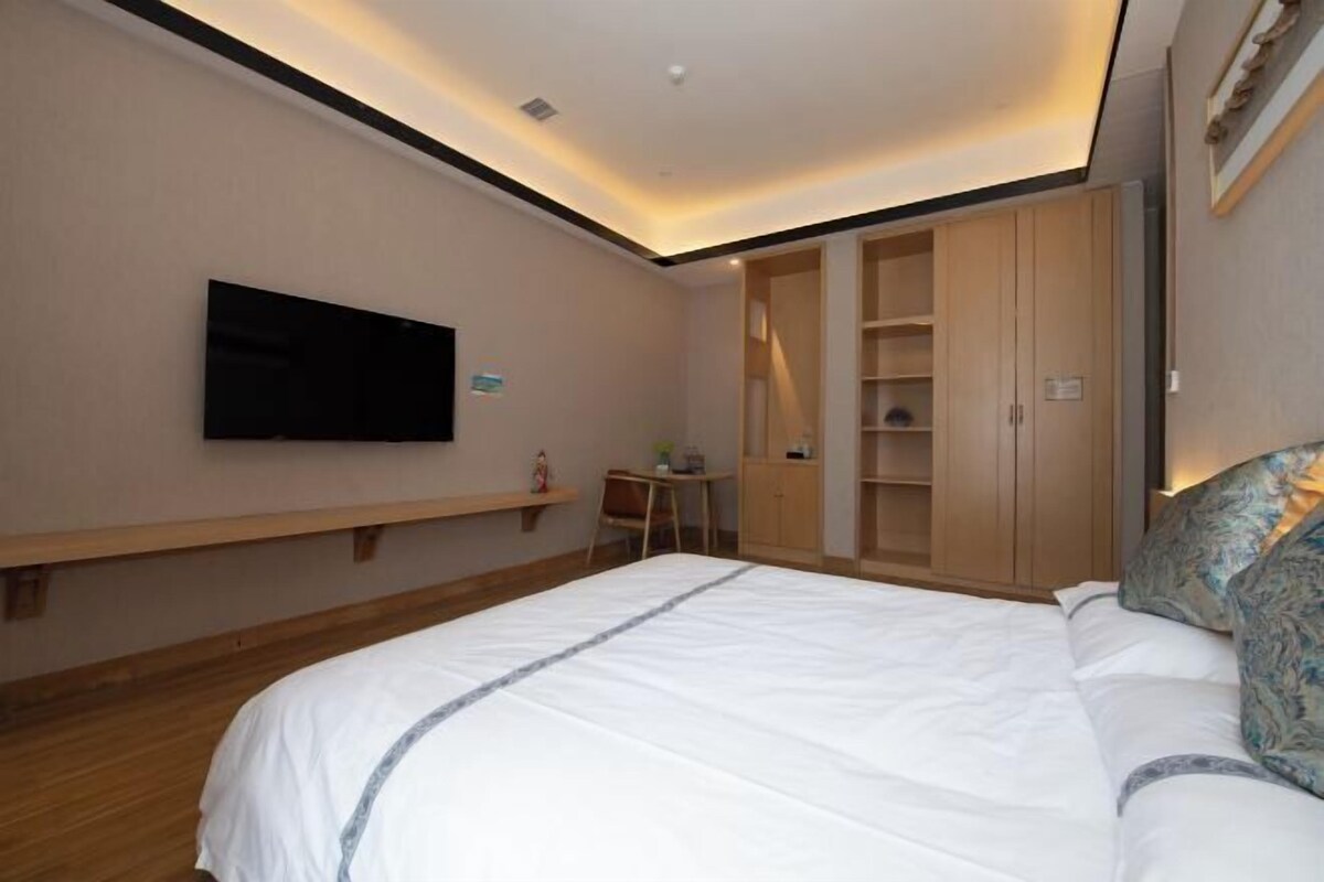 A hotel room with a large bed, a TV mounted on the wall, a wooden wardrobe, and a small table with a chair.