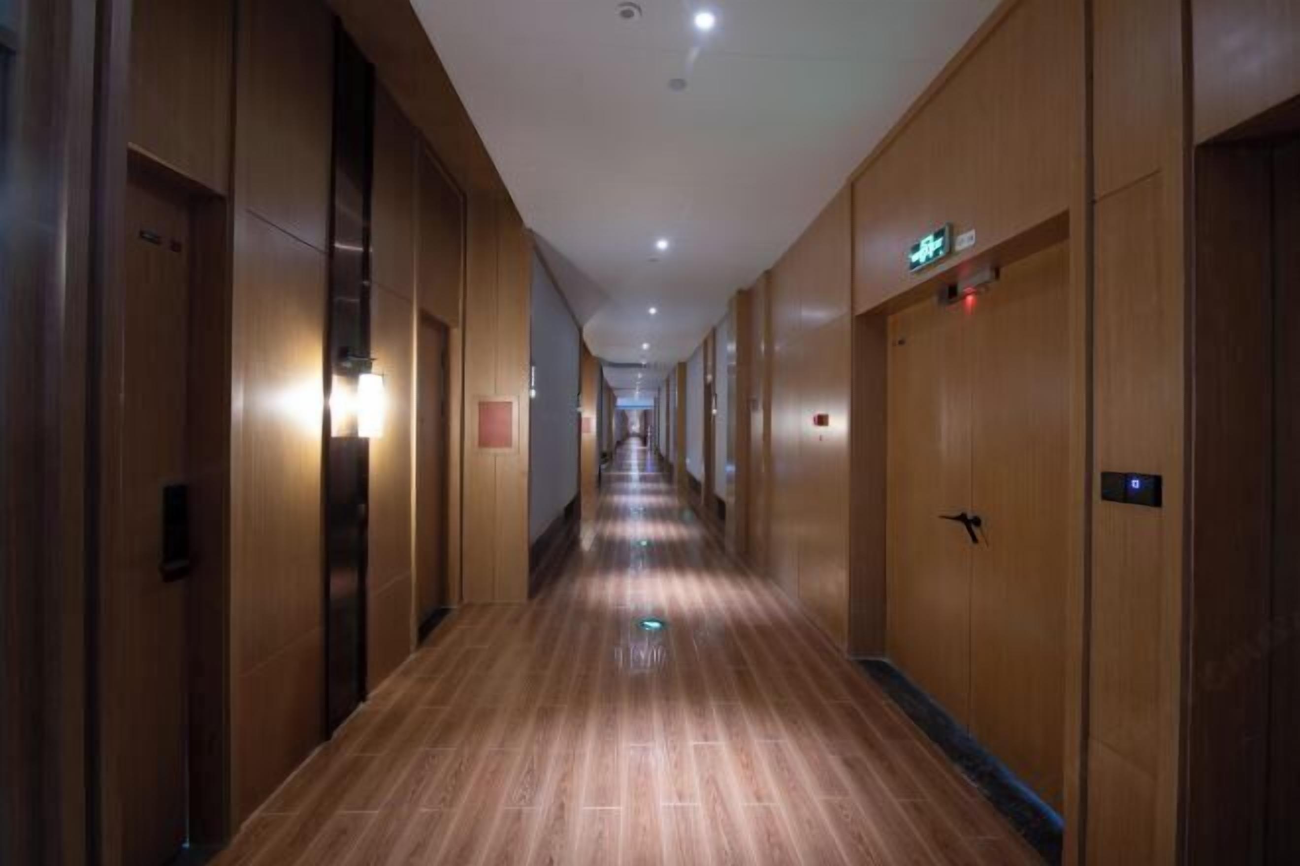 A long, narrow hotel corridor with wooden walls and doors, equipped with elevators and emergency exit signs.