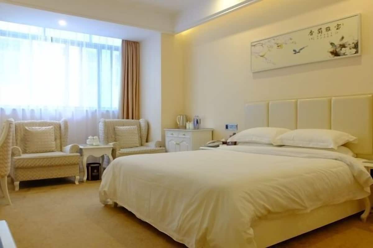 A hotel room with a large bed, two bedside tables, a chair, a nightstand, a wall-mounted artwork, and a window with curtains.