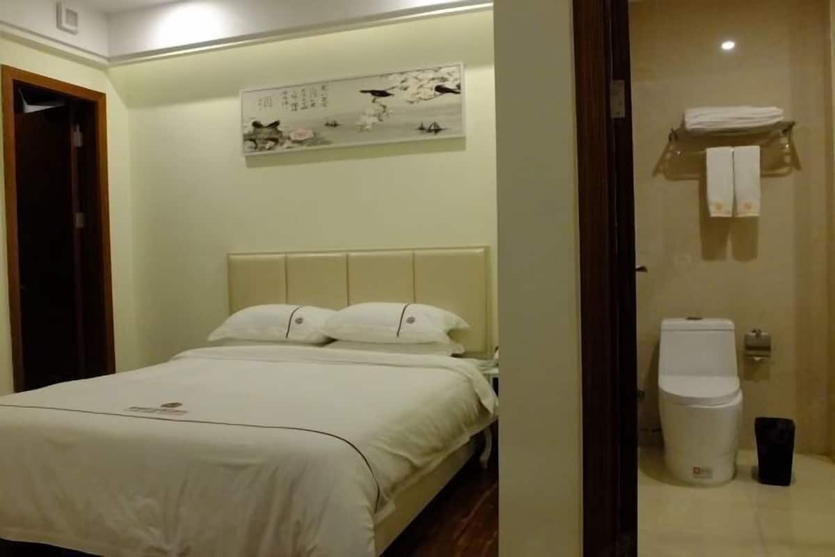 A hotel room with a bed, a wall-mounted artwork, a toilet, and a trash bin.