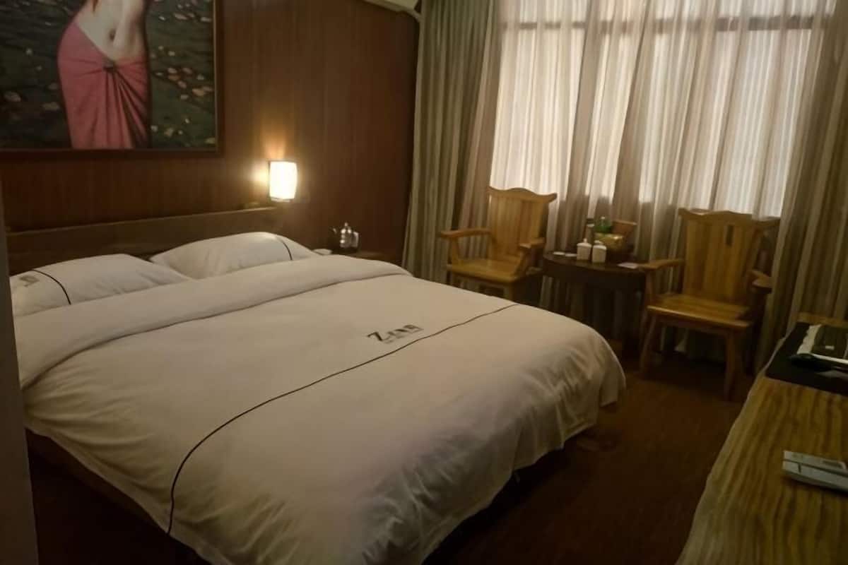 A hotel room with a large bed, two chairs, a small table, a painting on the wall, and a window with curtains.