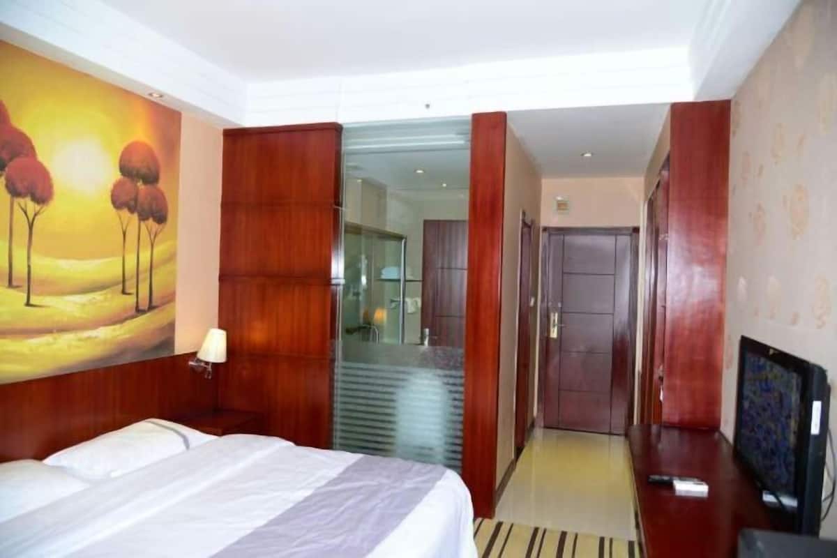 A hotel room with a bed, a TV, a painting on the wall, and a hallway leading to a bathroom.