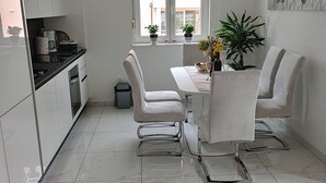 Une cuisine moderne avec des meubles blancs, une table à manger entourée de quatre chaises et une fenêtre équipée de stores.