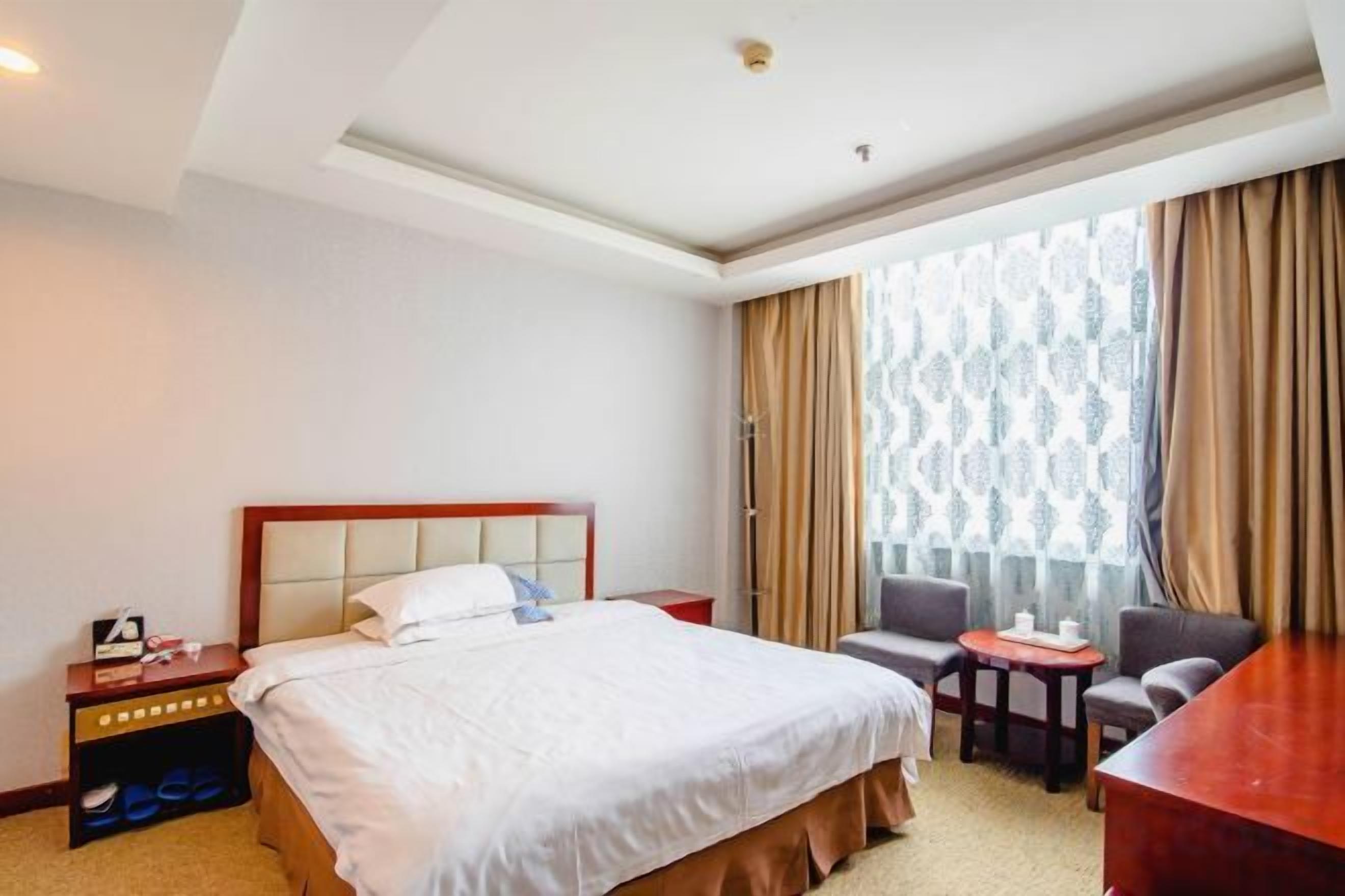 Quarto de hotel com cama, escrivaninha, cadeira, mesa pequena e janela com cortinas.