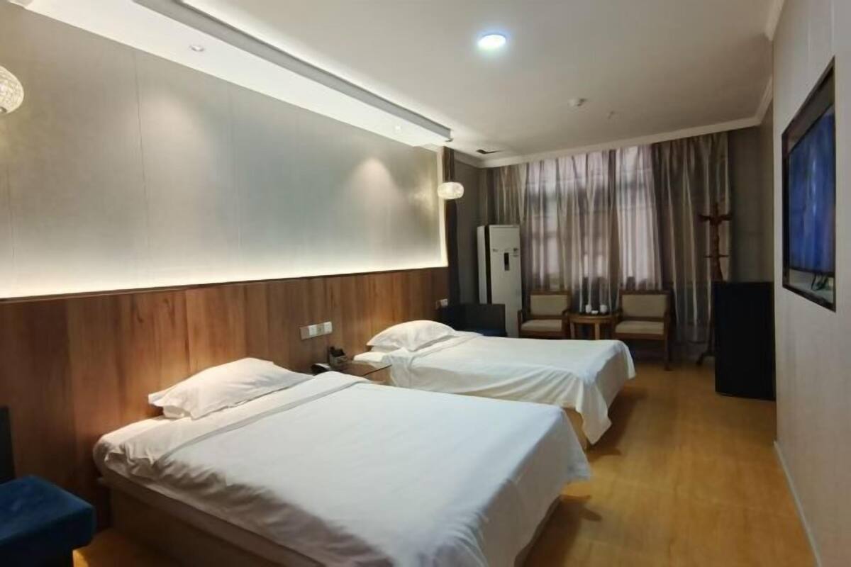 A hotel room with two beds, a wooden headboard, a desk, a chair, a TV, and a large window with curtains.