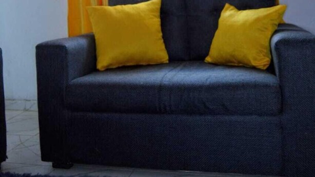 A grey sofa with two yellow cushions, a blue rug, and orange and white curtains with a window in the background. - Stay.Plus Mwanzo Apartment Eldoret (Eldoret)