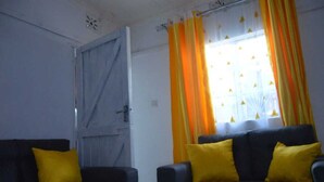 A room with two dark sofas, each with a yellow pillow, and a window with yellow and orange curtains. - Stay.Plus Mwanzo Apartment Eldoret (Eldoret)