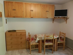 Huoneessa on puiset kaapit, jalustalla oleva televisio, pöytä tuoleineen ja pieni lipasto.