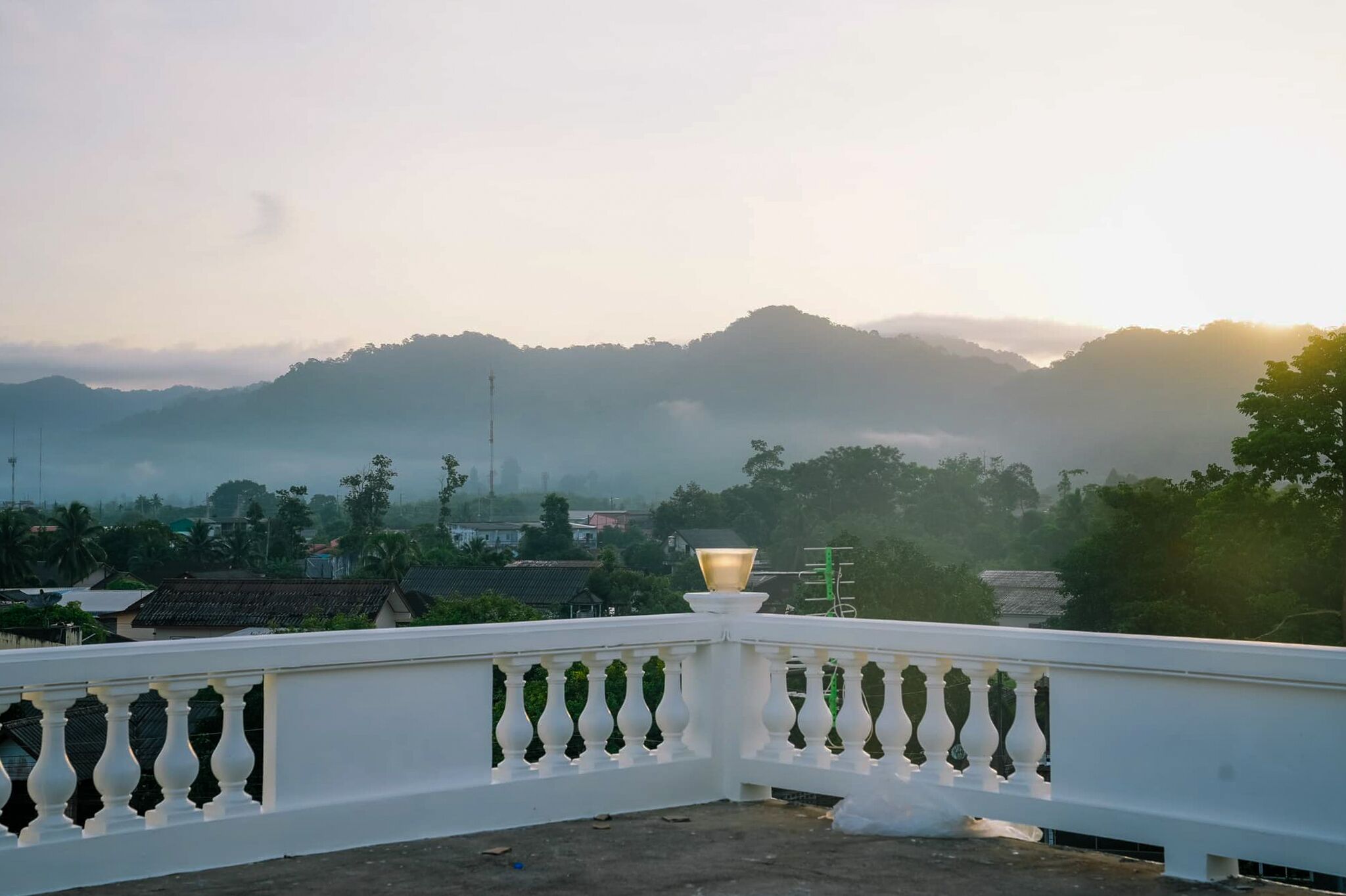 A balcony with a white balustrade overlooking a residential area with houses and trees, with mountains in the background.
