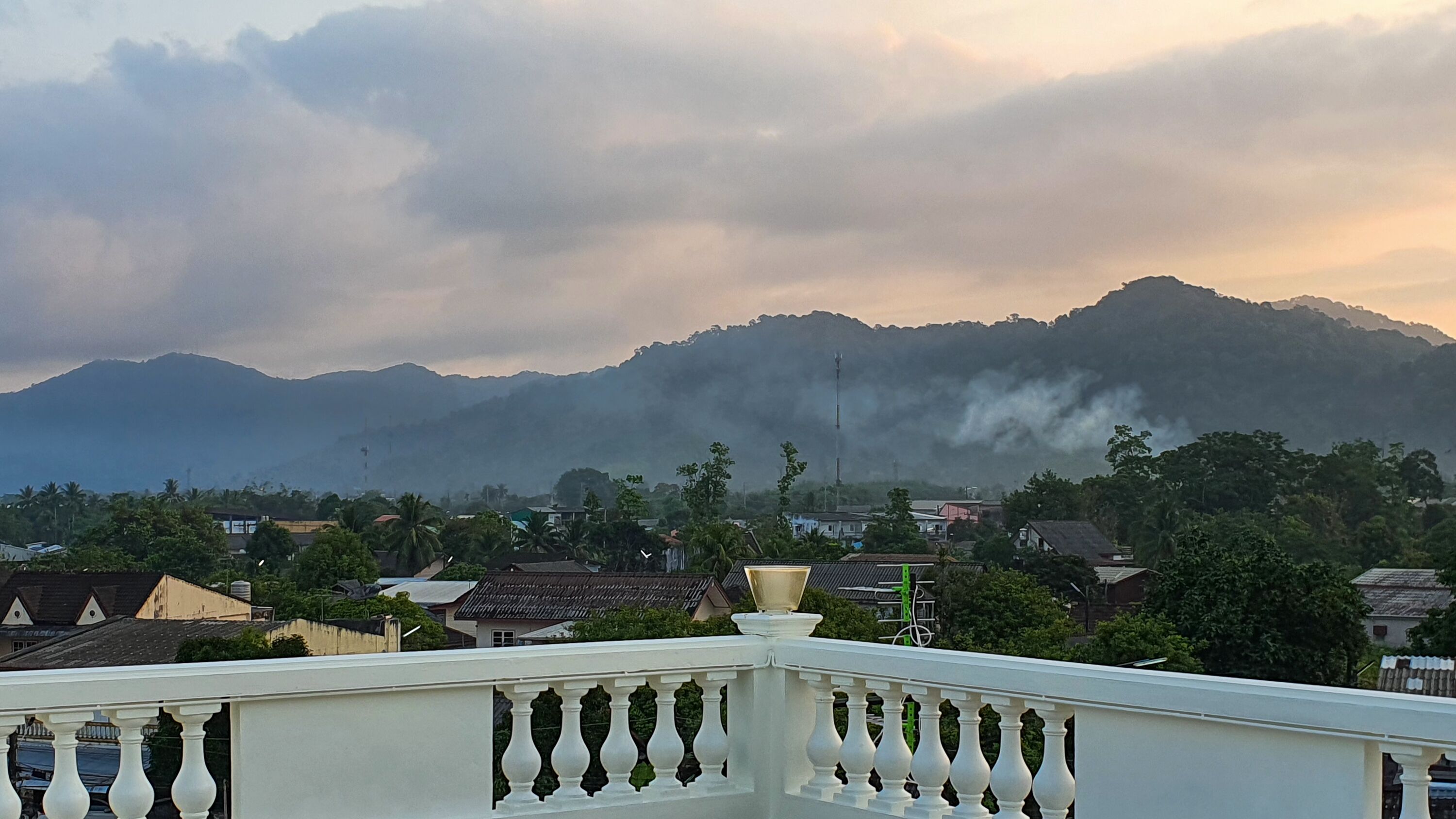 A balcony with a white balustrade overlooking a residential area with houses and a mountain in the background.