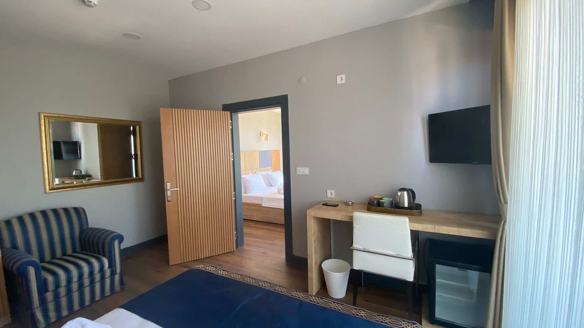 A hotel room with a bed, a desk, a chair, a TV, a mirror, and a window with curtains.