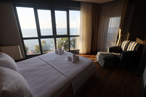 Habitación de hotel con un ventanal grande, cama con ropa blanca, un sillón a rayas y vista al mar.