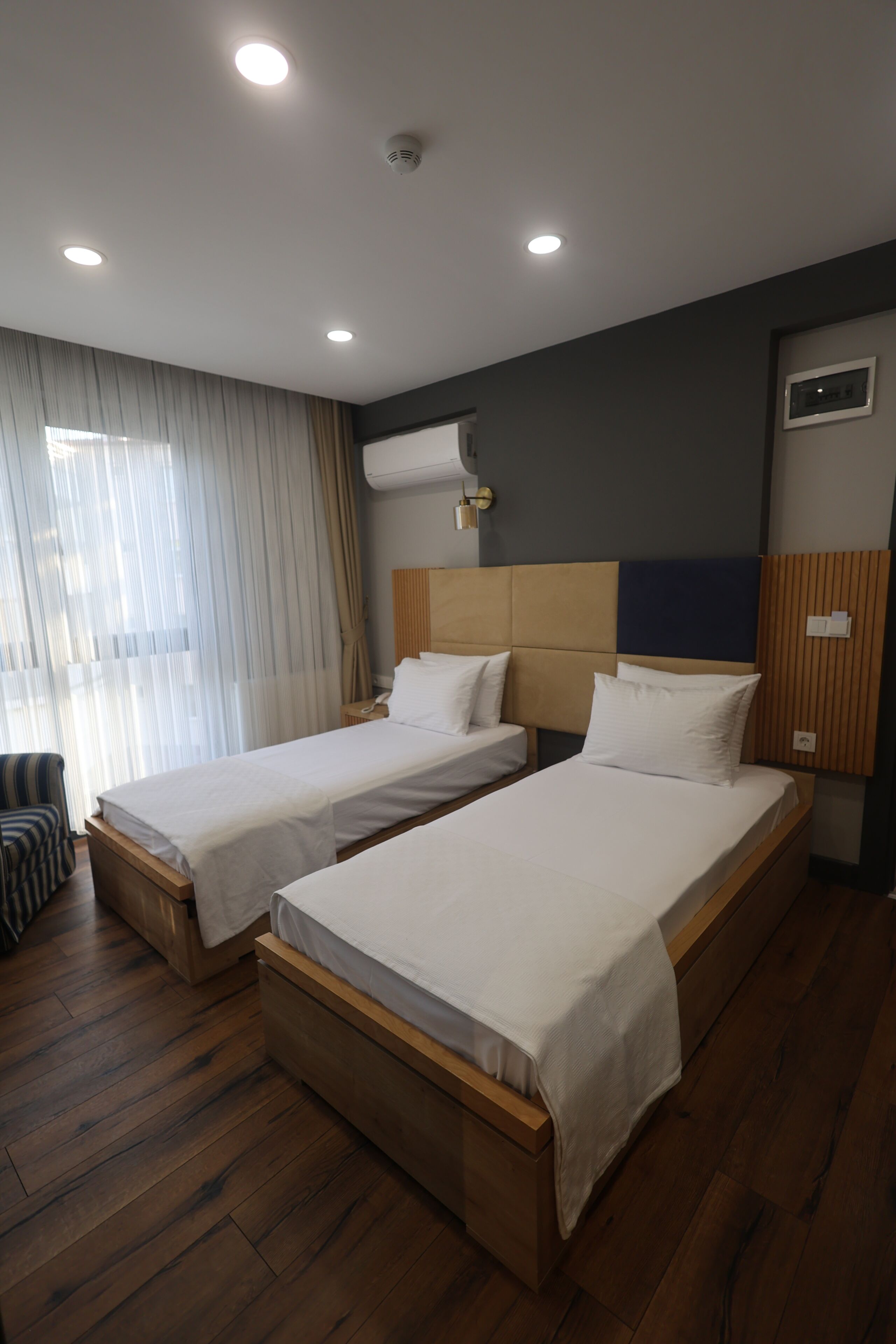 A hotel room with two beds, wooden flooring, a window with curtains, and a wall-mounted air conditioning unit.