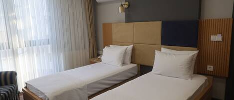 A hotel room with two beds, wooden flooring, a window with curtains, and a wall-mounted air conditioning unit.