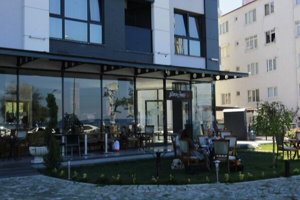 A modern building with a glass facade, outdoor seating area, and a sign that reads "Cafe". - Tasliman Otel (Cinarcik)