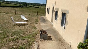A building with a concrete pathway leading to a door, two white lounge chairs, and a small garden area with a planter. - Peaceful farmhouse with valley view and terrace (Mazirat)