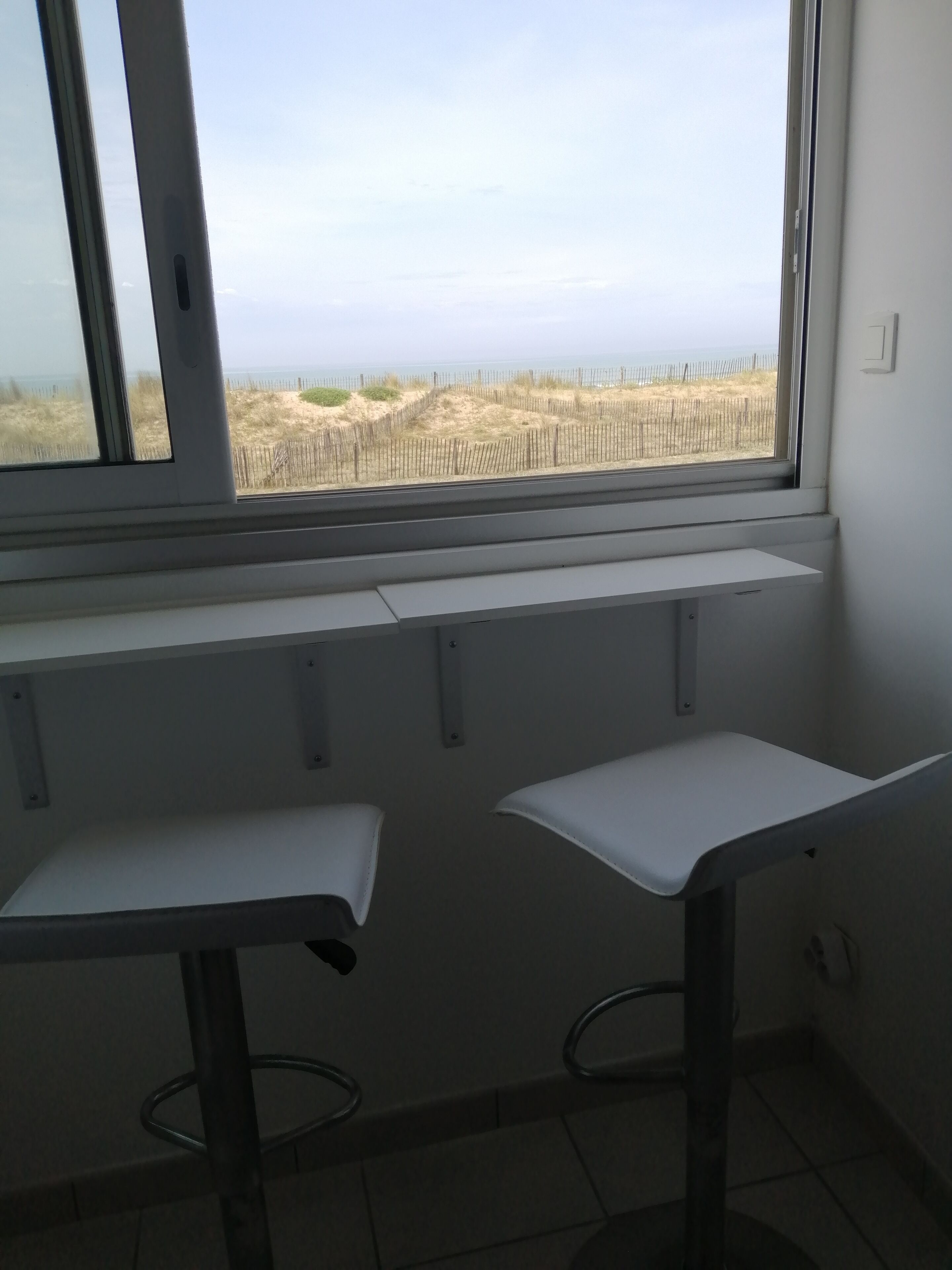 Een kamer met een groot raam dat uitkijkt op een zandstrand en een hek. Er staan twee witte tafels met metalen poten.