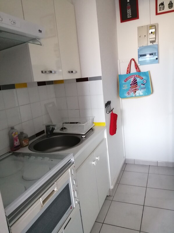 Cozinha com armários brancos, pia de aço inoxidável e escorredor de louça. Há produtos de limpeza no balcão e uma sacola azul pendurada na parede.