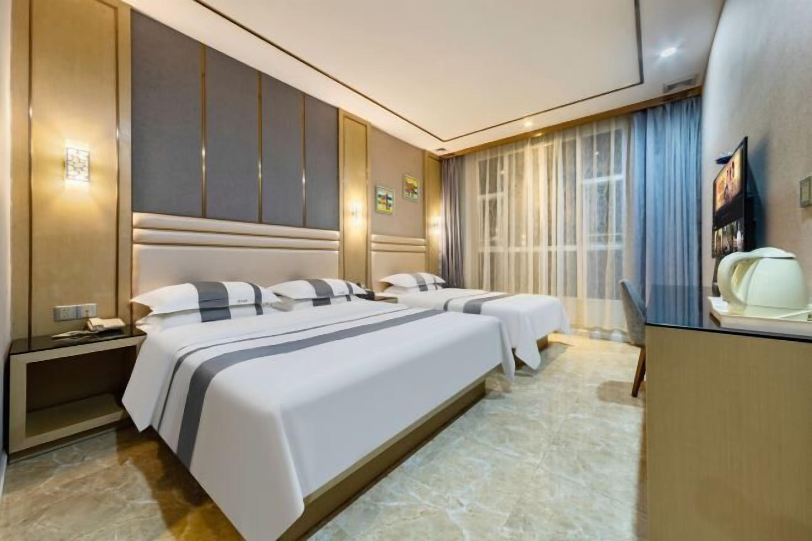 Quarto de hotel com duas camas, uma escrivaninha e uma janela grande com cortinas.