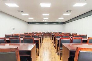 Een vergaderruimte met rijen tafels en stoelen, een houten vloer en verlichtingsarmaturen aan het plafond.