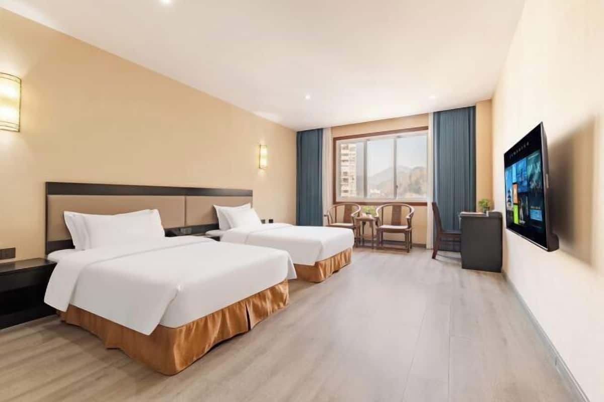 Ein Hotelzimmer mit zwei Betten, einem Schreibtisch, einem Stuhl, einem Fernseher und einem fensterbelaubten Fenster.
