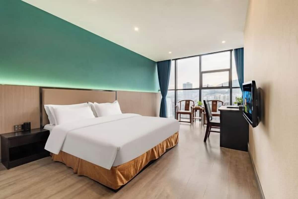 Ein Hotelzimmer mit einem großen Bett, einem Schreibtisch mit Stuhl, einem Fernseher und Blick auf die Stadt durch große Fenster.