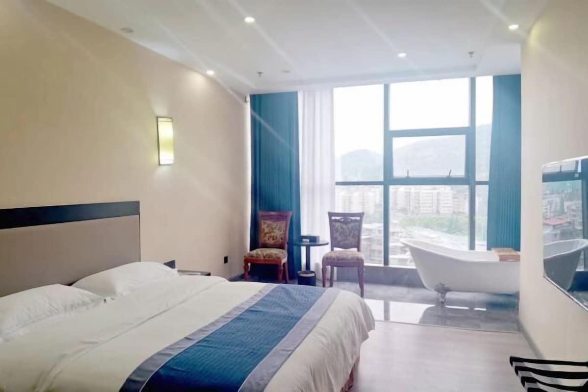 A hotel room with a large bed, a bathtub, and a view of the city through large windows.