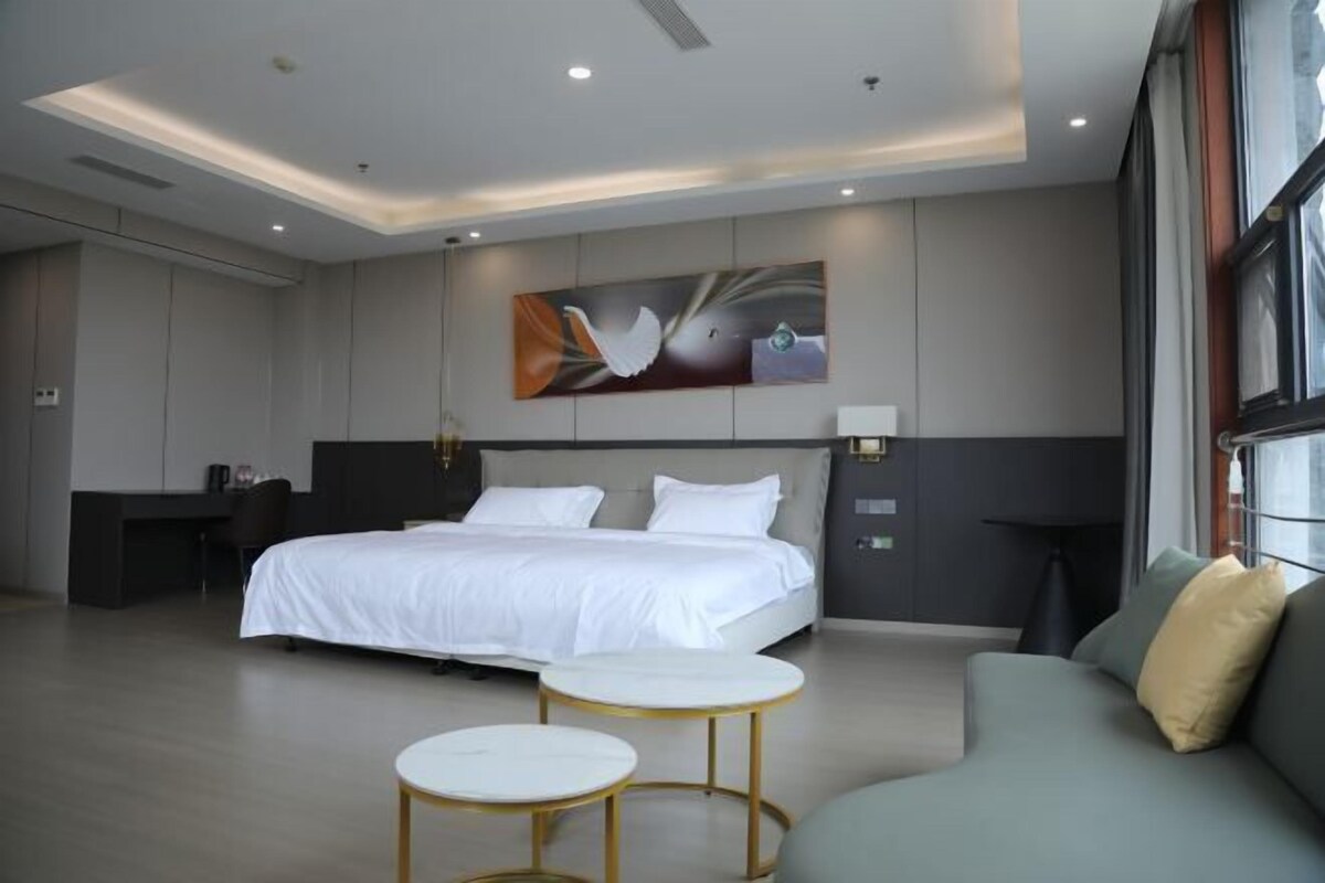 Quarto de hotel moderno com uma cama grande, um sofá, duas mesinhas e um quadro na parede.