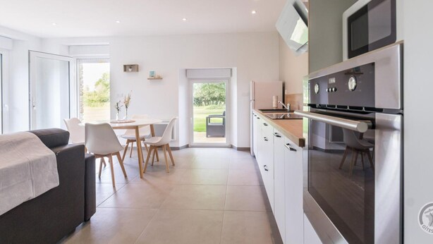 A modern kitchen with stainless steel appliances, a dining area with white chairs, and a view of greenery outside. - Single-storey house in Anneville sur Mer with private garden (Gouville-sur-Mer)