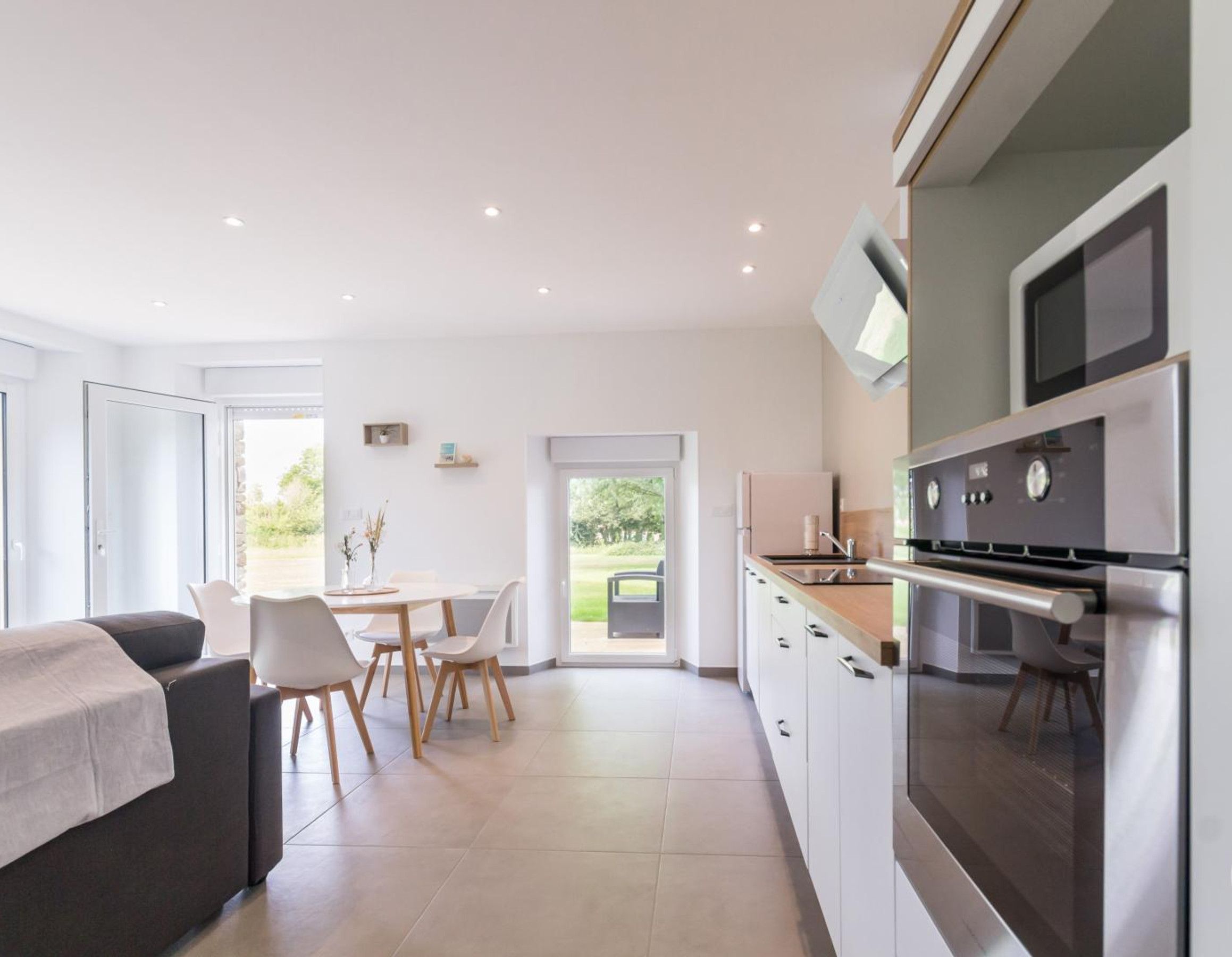 Une cuisine moderne équipée d’appareils électroménagers en acier inoxydable, un coin repas avec des chaises blanches et une vue sur la verdure environnante.