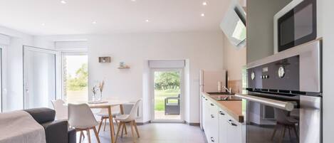 Une cuisine moderne équipée d’appareils électroménagers en acier inoxydable, un coin repas avec des chaises blanches et une vue sur la verdure environnante.