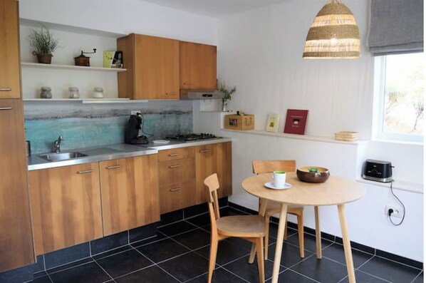 A kitchen with wooden cabinets, a black tiled floor, a round wooden table with two chairs, a hanging light fixture, and a window with a view. - Lovely detached cocoon in green property-terrace/garden (Barjols)