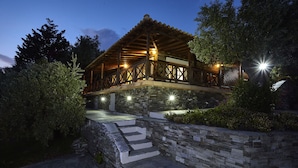 A two-story wooden structure with a stone base, surrounded by greenery and a stone pathway leading to the entrance. - Skiathos Ira Hotel (Skiathos)