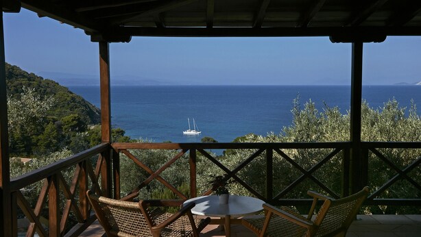 A wooden deck with wicker chairs and a small table, overlooking a coastal view with a sailboat on the water. - Skiathos Ira Hotel (Skiathos)
