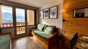 Une pièce chaleureuse avec un plancher en bois, un canapé vert, une table à manger avec de l’assiette, et une vue sur des montagnes enneigées par de grandes fenêtres.
