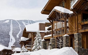 Een houten lodge met stenen zuilen, een skipiste op de achtergrond en met sneeuw bedekte bomen.