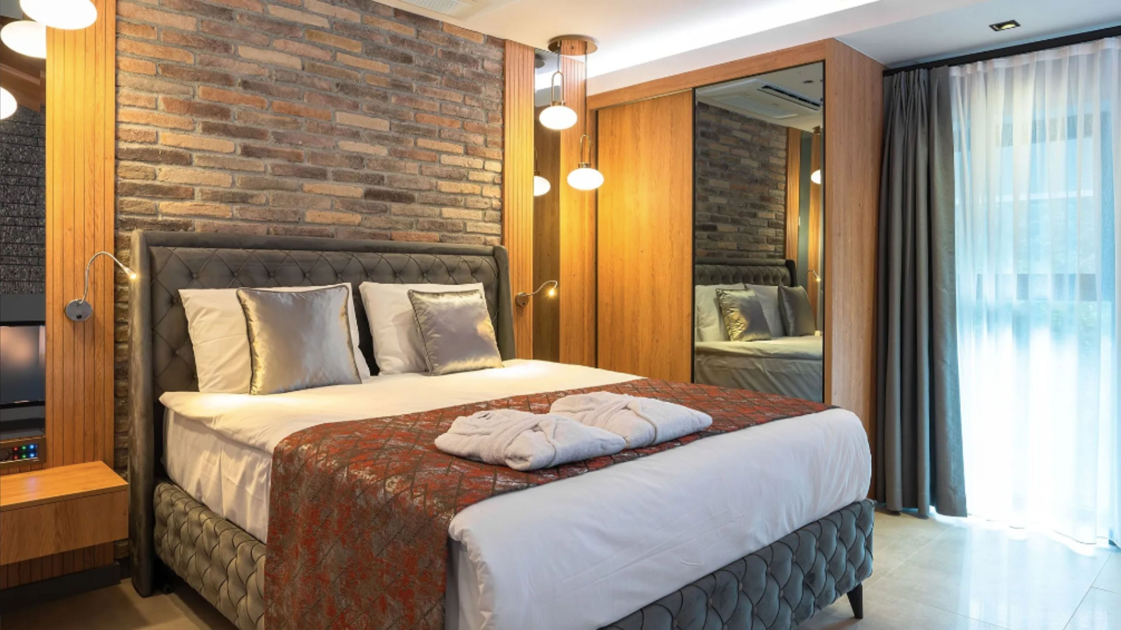 Une chambre d’hôtel moderne avec un mur en briques, un grand lit recouvert d’une courtepointe à motifs rouges et blancs, une table de chevet en bois et un grand miroir qui reflète la pièce.