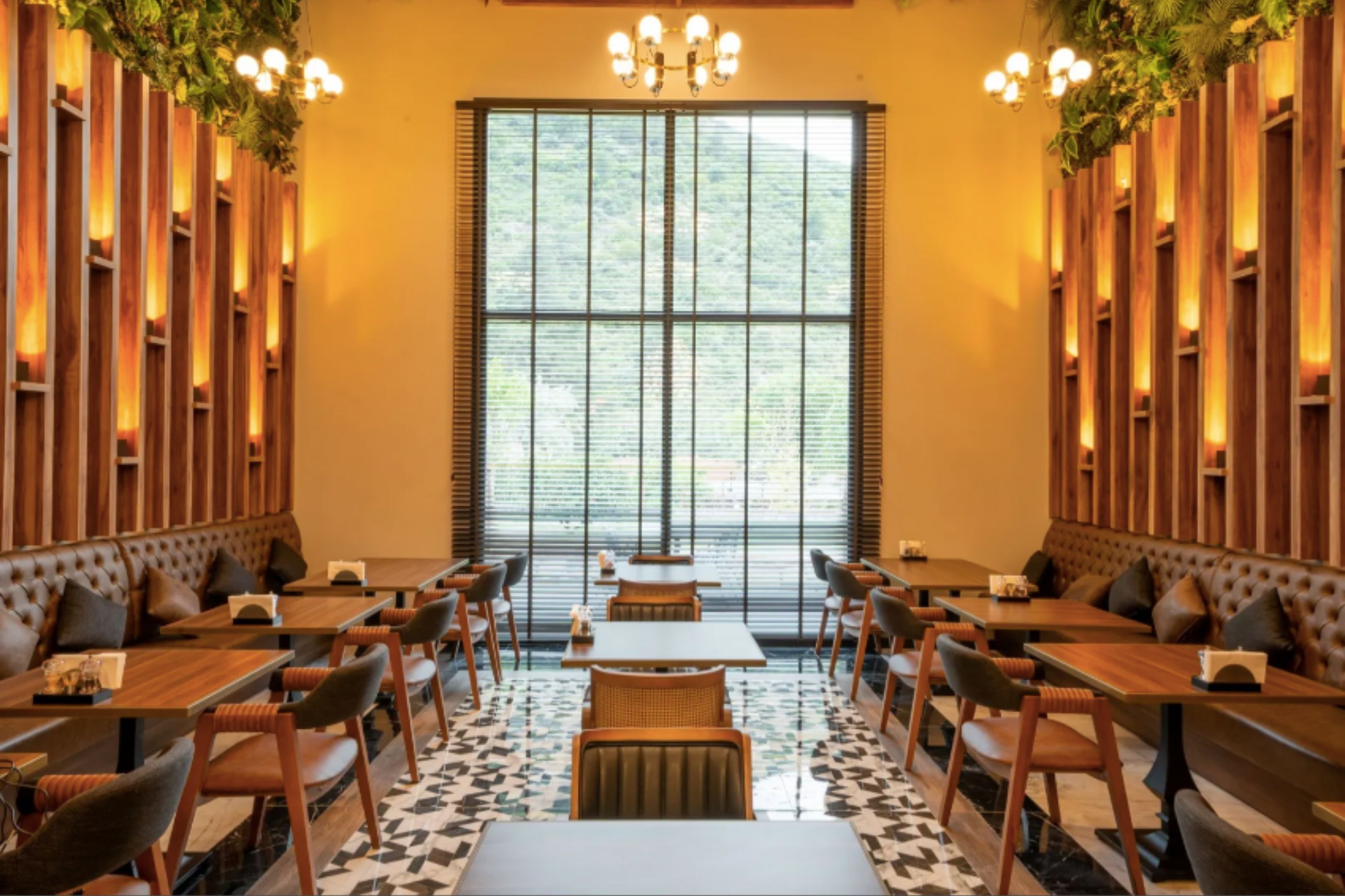 Un restaurante con paneles de madera, ventanas grandes y una araña. Mesas y sillas dispuestas para comer.