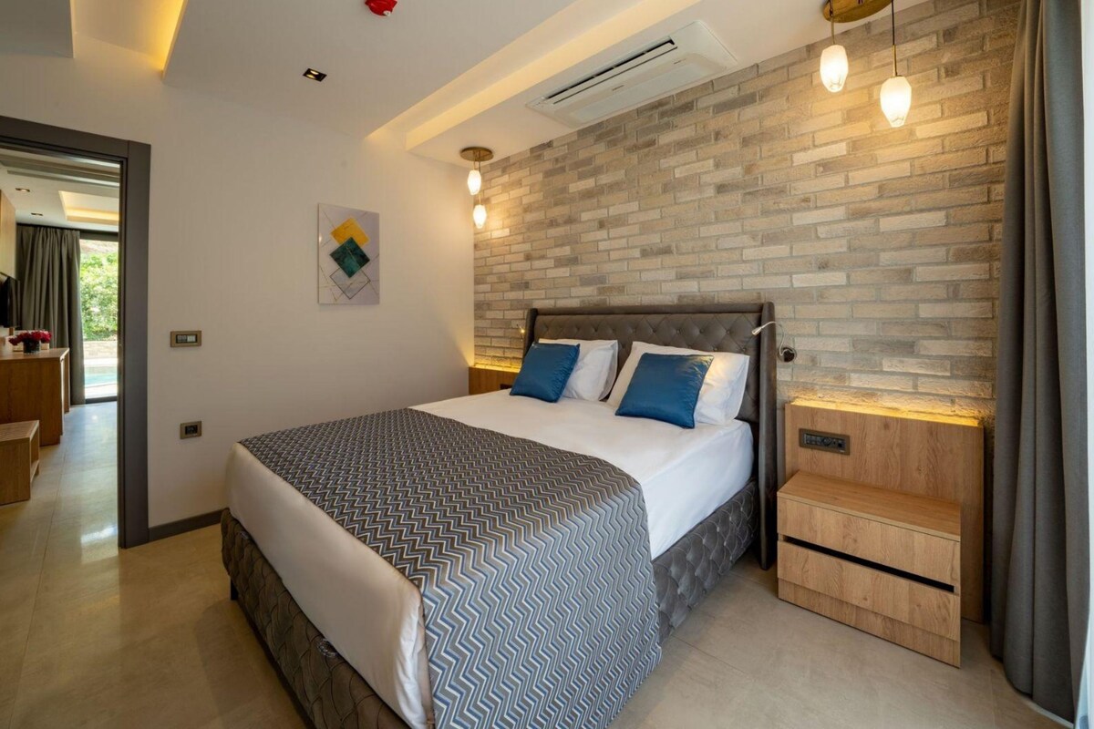 A modern bedroom with a brick-patterned headboard, a bed with a geometric-patterned bedspread, two bedside tables with lamps, and a small wooden nightstand.