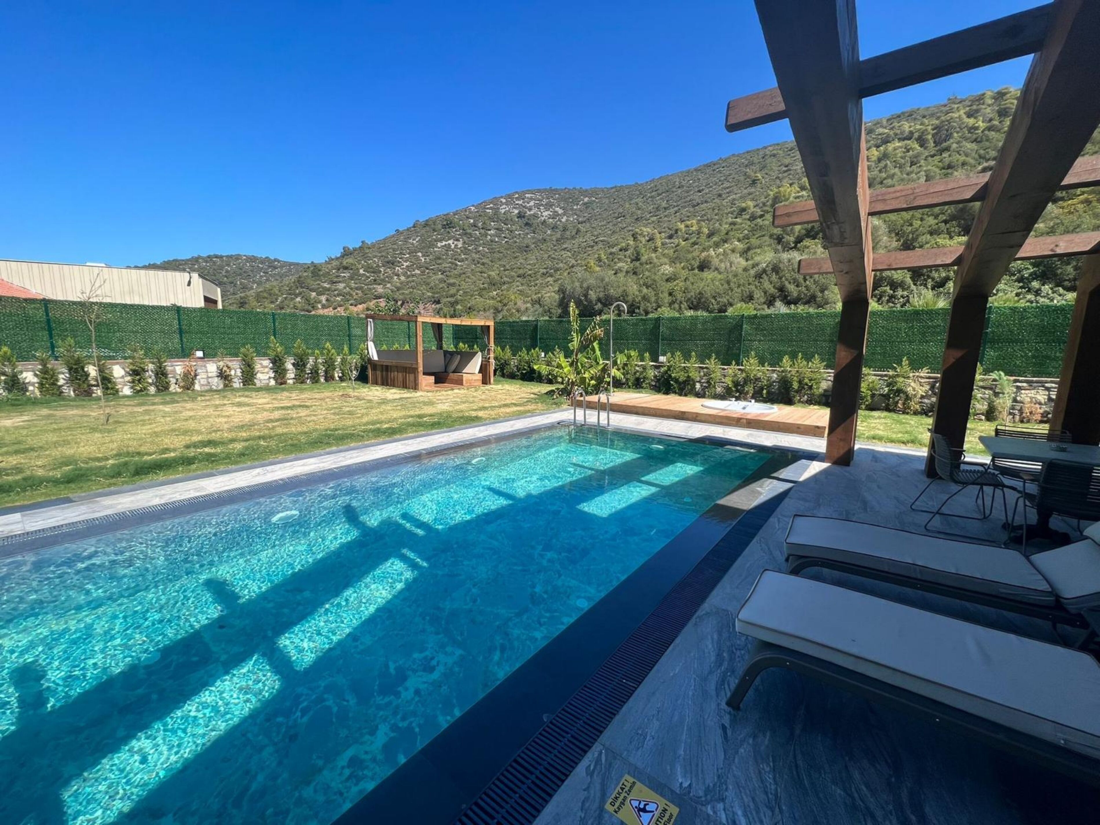 Una piscina con muelle de madera y sillones de descanso, rodeada de una cerca verde y montañas al fondo.