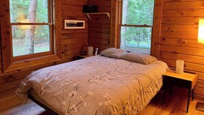 A wooden cabin bedroom with a bed, two windows, a bedside table, and a framed picture on the wall.