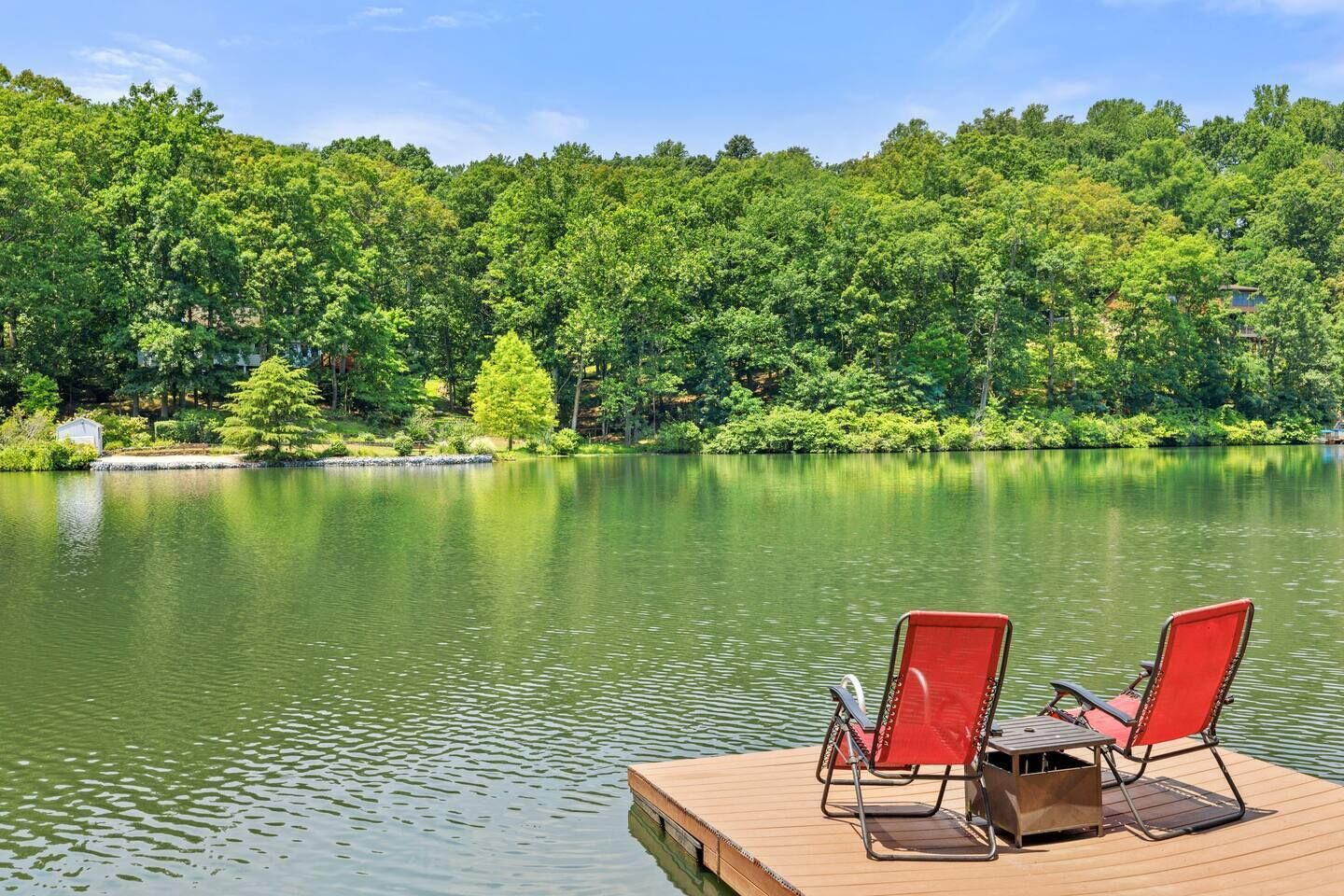 A wooden dock with two red chairs and a table, overlooking a calm lake surrounded by dense green forest.