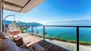 Un balcón con una silla colgante, piso de madera y vista a un mar tranquilo y montañas lejanas.