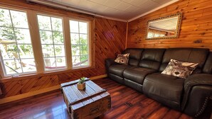 A cozy living room with a leather sofa, wooden walls, a wooden coffee table, and a large window with a view of outdoor furniture.