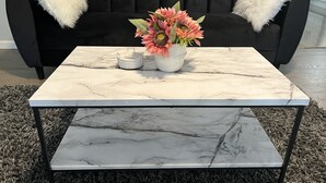A black sofa with white fluffy pillows, a white vase with pink flowers, and a marble coffee table with a metal frame.