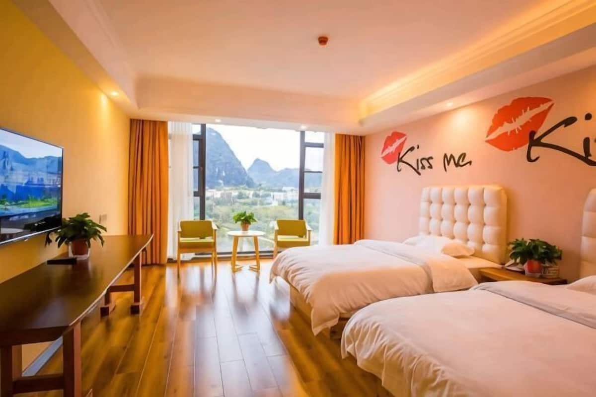 Habitación de hotel con dos camas, televisor, escritorio, silla, mesita y ventana con vista a la montaña.
