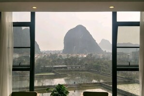 A room with a large window offering a view of a mountainous landscape and a body of water. - Tao Flute Hotel (Guilin)
