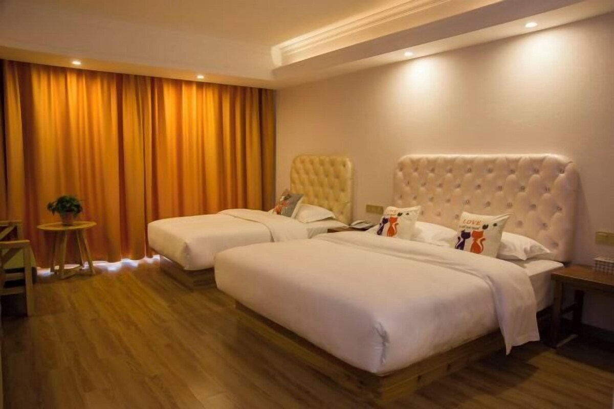 A hotel room with two beds, a wooden floor, a small round table with a plant, and a large window with curtains.