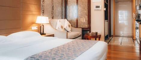 Una habitación de hotel con una cama, una mesita de noche, una lámpara, una silla, una pequeña mesa y una puerta que da a un pasillo.
