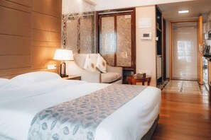A hotel room with a bed, a nightstand, a lamp, a chair, a small table, and a door leading to a hallway.